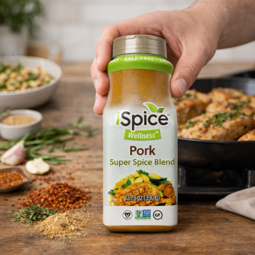 Hand holding a bottle of iSpice Wellness Pork Super Spice Blend on a wooden table with ingredients in the background.